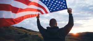 A person stands outdoors at sunset, holding up a large American flag with hills and a cloudy sky in the background, symbolizing hope—much like an Arlington temporary protected status lawyer helping clients achieve their dreams.