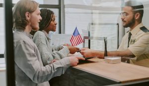 Two people hand documents to a uniformed employee behind a glass divider at an office desk with an American flag, assisted by a Fort Worth immigration attorney.