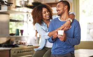 A woman smiles and hugs a man from behind in a kitchen while he holds a white mug, both appearing happy and relaxed—just like couples assisted by Grand Prairie K-1 fiancé visa attorneys.