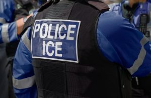 A close-up of a law enforcement officer wearing a vest labeled "POLICE ICE," when detained by ICE consult with deportation defense lawyer Katy.