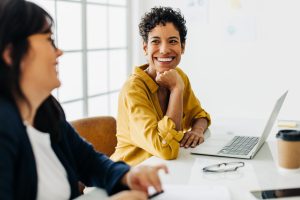 Two women sit at a desk with a laptop and coffee cup, engaged in conversation; one woman, a Katy VAWA lawyer, is smiling brightly.