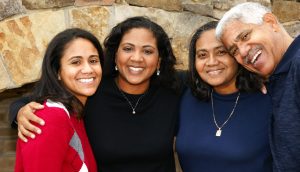 Four adults stand closely together, smiling in front of a stone wall. Three women and one man pose with their arms around each other, representing the warmth and unity found in family based immigration in The Woodlands.