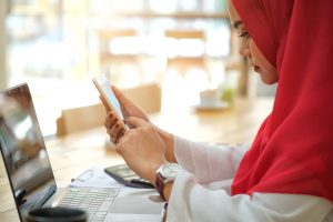 A woman in a red hijab sits at a desk using a smartphone, with an open laptop and notebook in front of her, possibly researching the best asylum attorney in Houston.