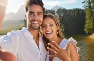 A couple smiles at the camera outdoors by a lake; the woman shows an engagement ring on her hand, celebrating their love and recent k-1 visa Houston approval.