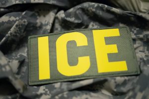 A yellow “ICE” patch on a grey digital camouflage uniform worn by an immigration detention attorney in Houston.